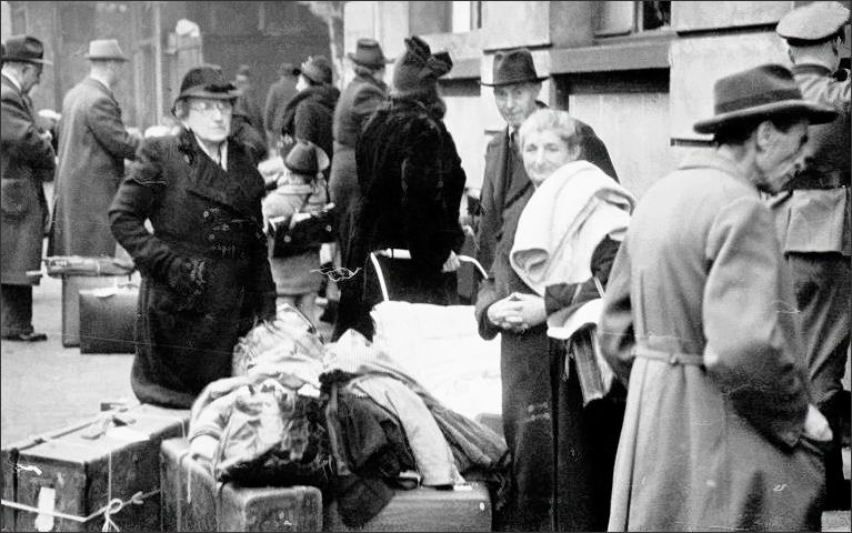 Das Foto zeigt Jüdinnen und Juden, die auf die Durchsuchung ihres Gepäcks und den Zwangsumtausch der ihnen verbliebenen Barmittel warten. Elsa Löb (mit Brille), die an der Kamera vorbei blickt, wurde 1942 in Auschwitz-Birkenau ermordet. Ludwig Pinkus (mittig), der in die Kamera schaut, überlebte, starb aber schon 1947 in Aix-les-Bains. Über die Frau mit der Decke über der Schulter ist nichts bekannt. (Foto eines unbekannten Fotografen/einer unbekannten Fotografin | Quelle: Marchivum, Bildsammlung, KF013142)